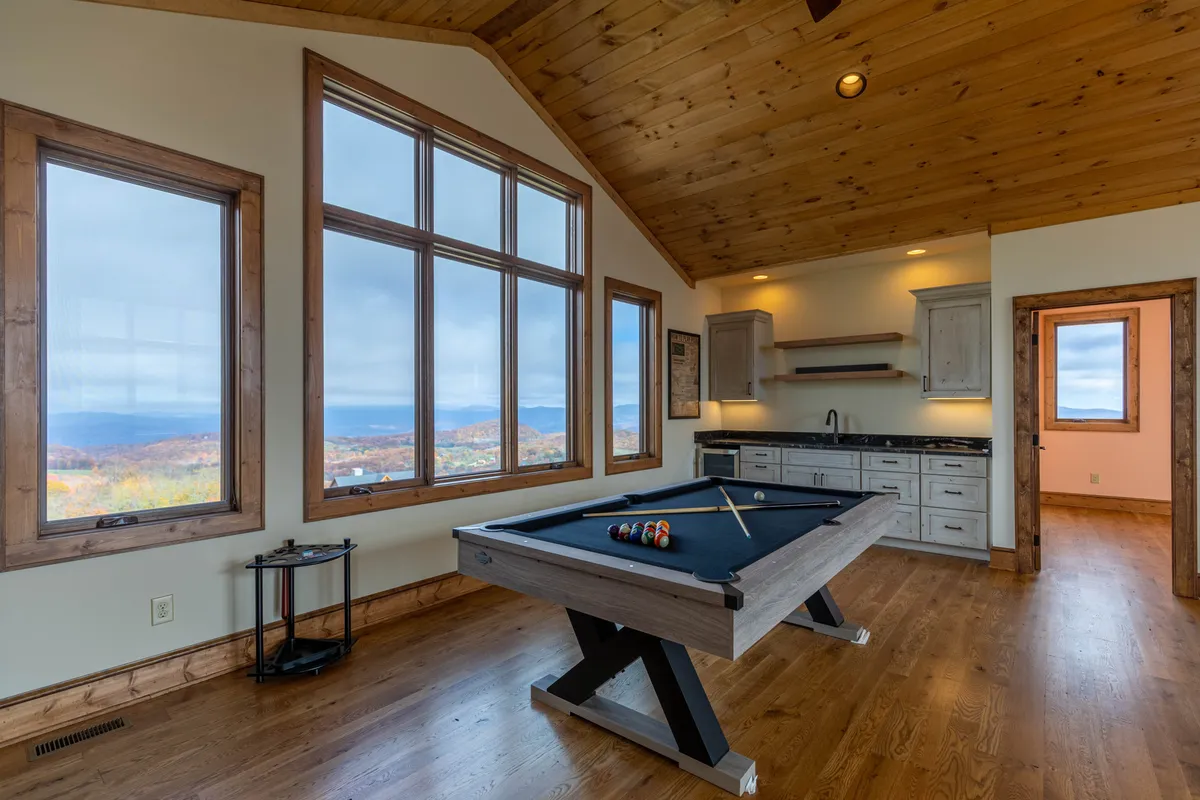 This image features a spacious loft area centered around a pool table, with large windows that frame scenic mountain views. The space includes a wooden ceiling, which adds warmth and a rustic touch. The bright natural light streaming through the windows creates an inviting atmosphere for games and relaxation.