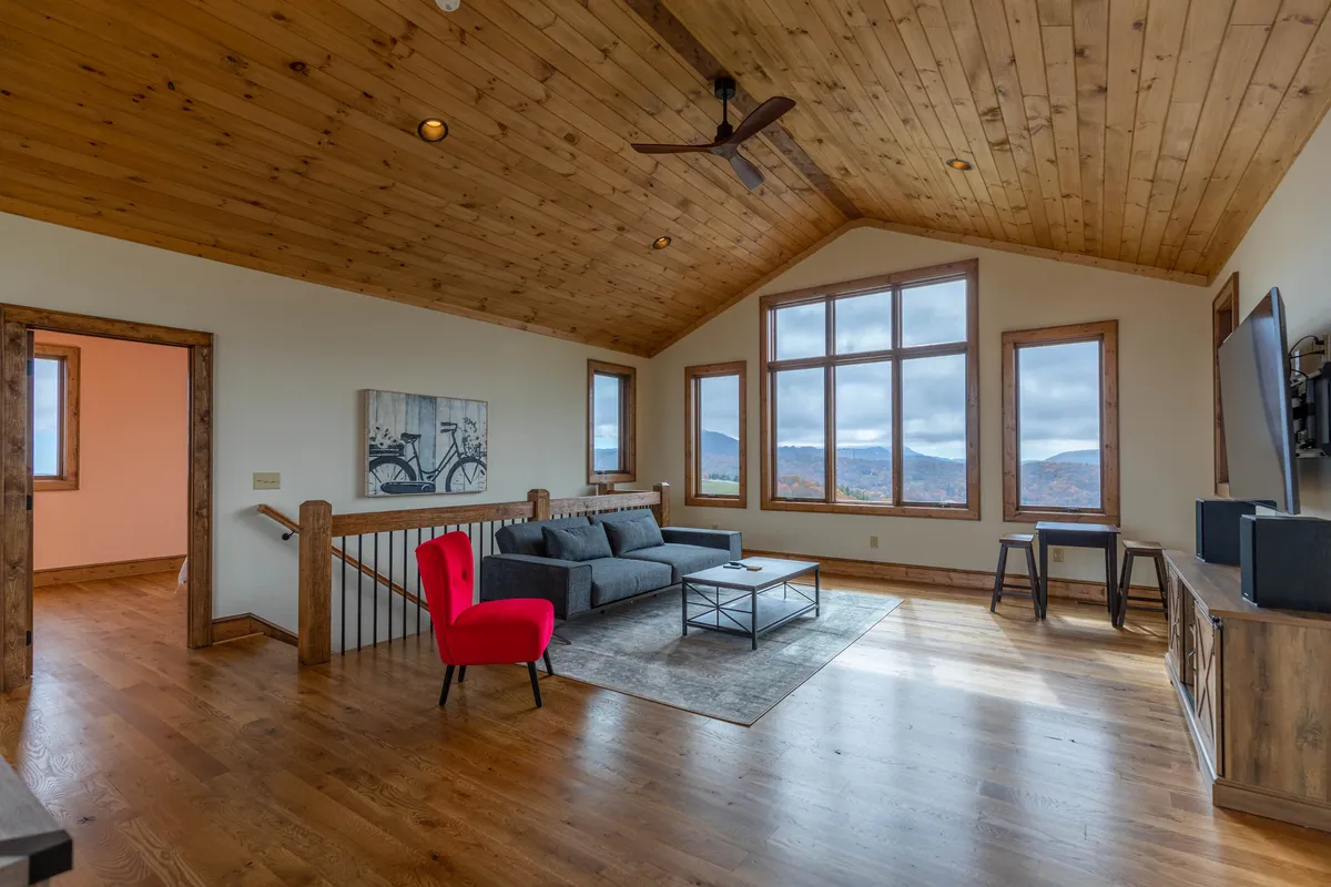 A spacious, inviting living area featuring a cozy red accent chair, a sofa arrangement designed for socializing, and large windows that open up to breathtaking mountain vistas. The warm wooden ceiling adds to the cozy, celebratory ambiance.
