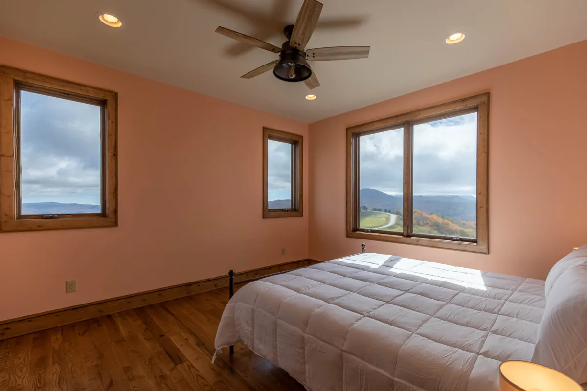 A serene bedroom with a plush bed is surrounded by windows that offer breathtaking vistas of the surrounding mountains. The ceiling fan and airy layout enhance the tranquil, nature-infused vibe, inviting guests to relax and take in the sights and sounds of the High Country.