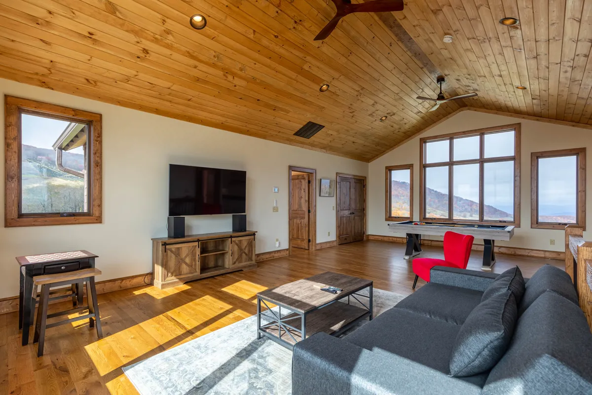 A warm, inviting lounge area with a large window showcasing sweeping mountain views, a red accent chair, dark wood furniture, and a soft sofa. The scene suggests shared moments—playing games, watching TV, or simply soaking in the sunset with loved ones.