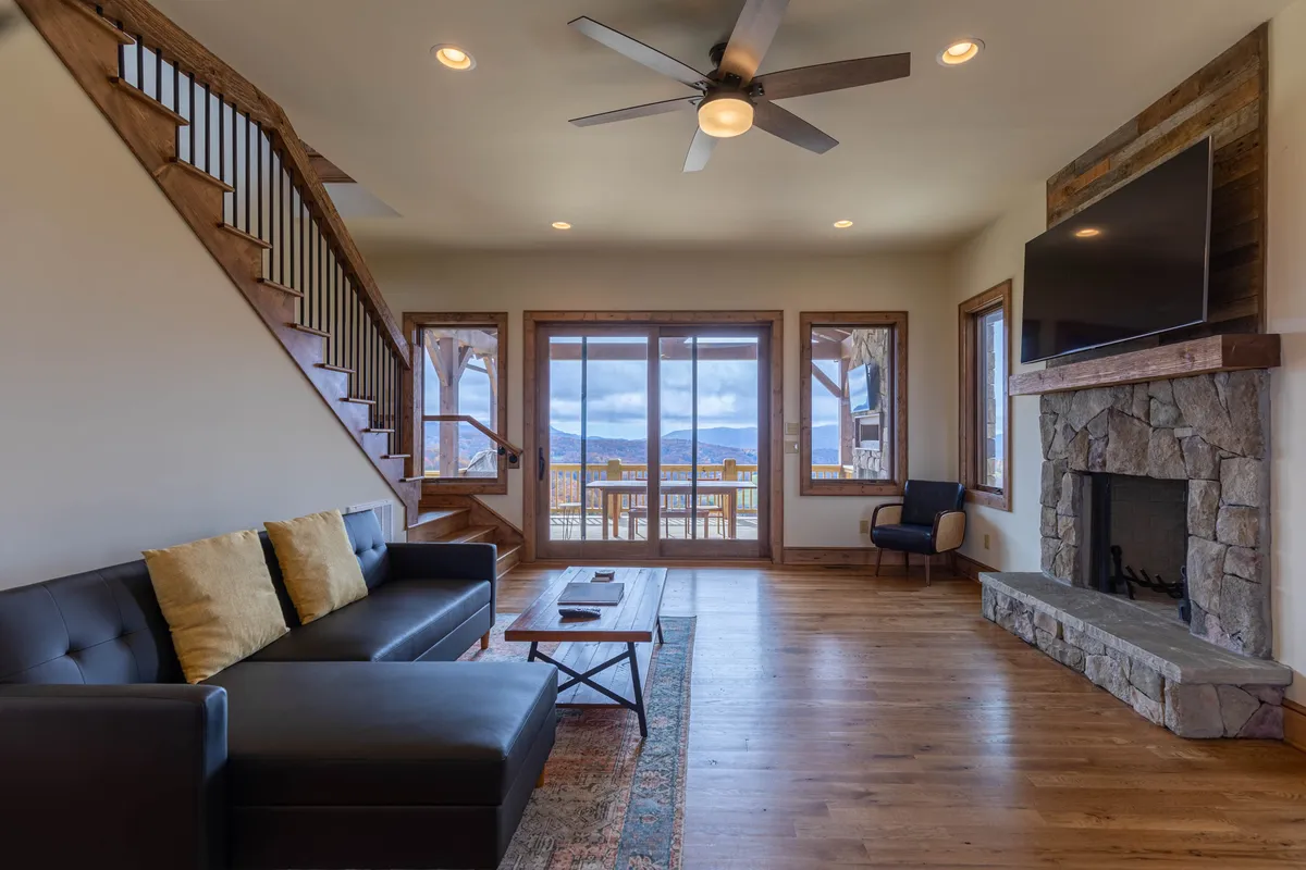 This image features a comfortable living area with a plush sofa, a striking fireplace, and large windows that flood the space with natural light and offer expansive mountain views. The staircase hints at the multi-level layout, and the overall setting exudes a welcoming, communal atmosphere—ideal for group relaxation after a day of local adventures.