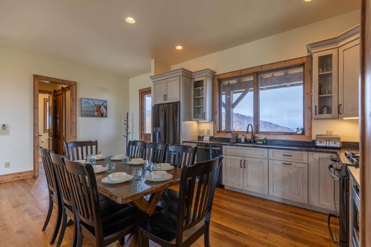 The dining table is elegantly set, with stylish kitchen cabinetry in the background and expansive windows framing mountain views. The atmosphere is cozy and communal, inviting guests to partake in celebratory meals and shared traditions in a space infused with natural light.