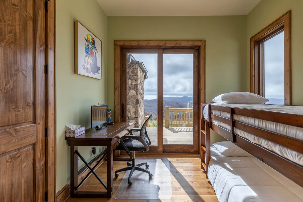 A cozy bunk bed sits alongside a desk equipped with a computer, positioned near large windows that frame sweeping views of the mountains beyond. Natural light floods the space, inviting guests to soak in the scenery or glimpse the stars at night. The room seamlessly connects indoor comfort with the outdoors, highlighting both work and relaxation zones.
