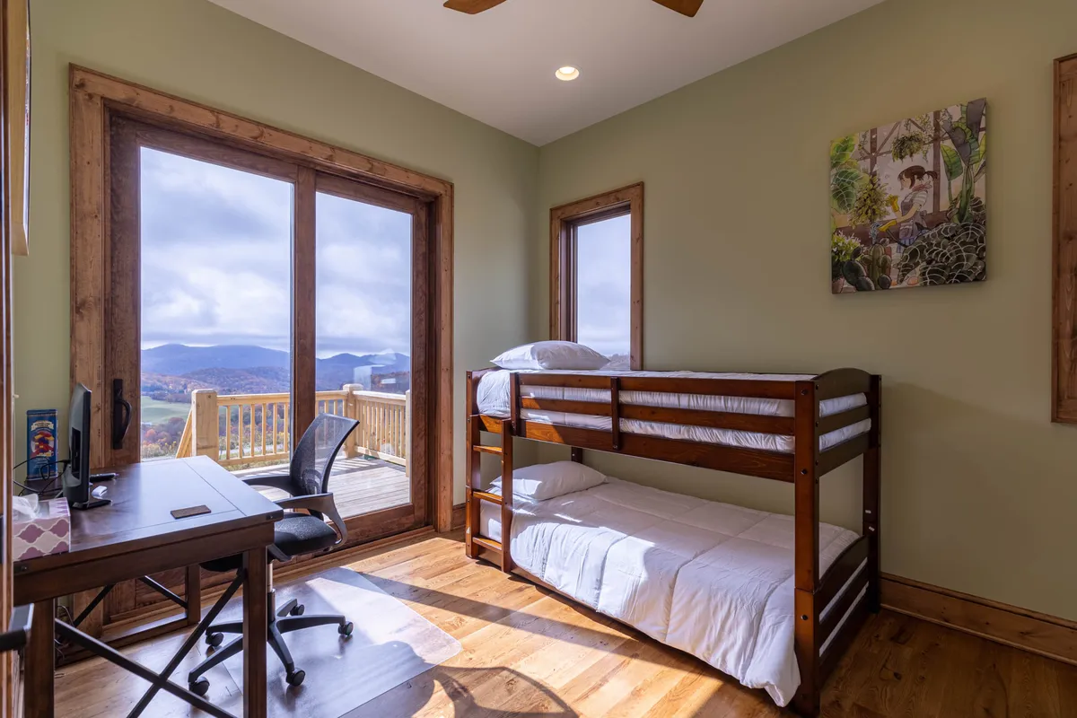 A cozy bunk room featuring built-in bunk beds, a desk with a chair positioned by a large window, and charming wall art. The space is bright and welcoming, ideal for children or adventurous guests, with views of the mountains visible through the windows.