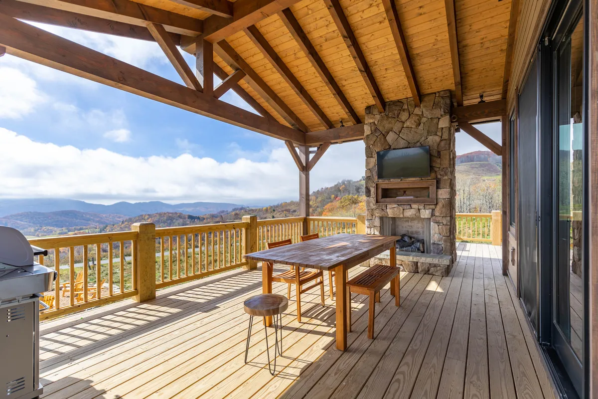 This image showcases a spacious wooden deck featuring a large outdoor dining table, a striking stone fireplace, and expansive mountain views. The setup is perfect for communal meals, cozy fireside gatherings, and enjoying the natural surroundings. The scene conveys a blend of comfort and outdoor luxury, inviting guests to relax and dine al fresco while taking in the views.