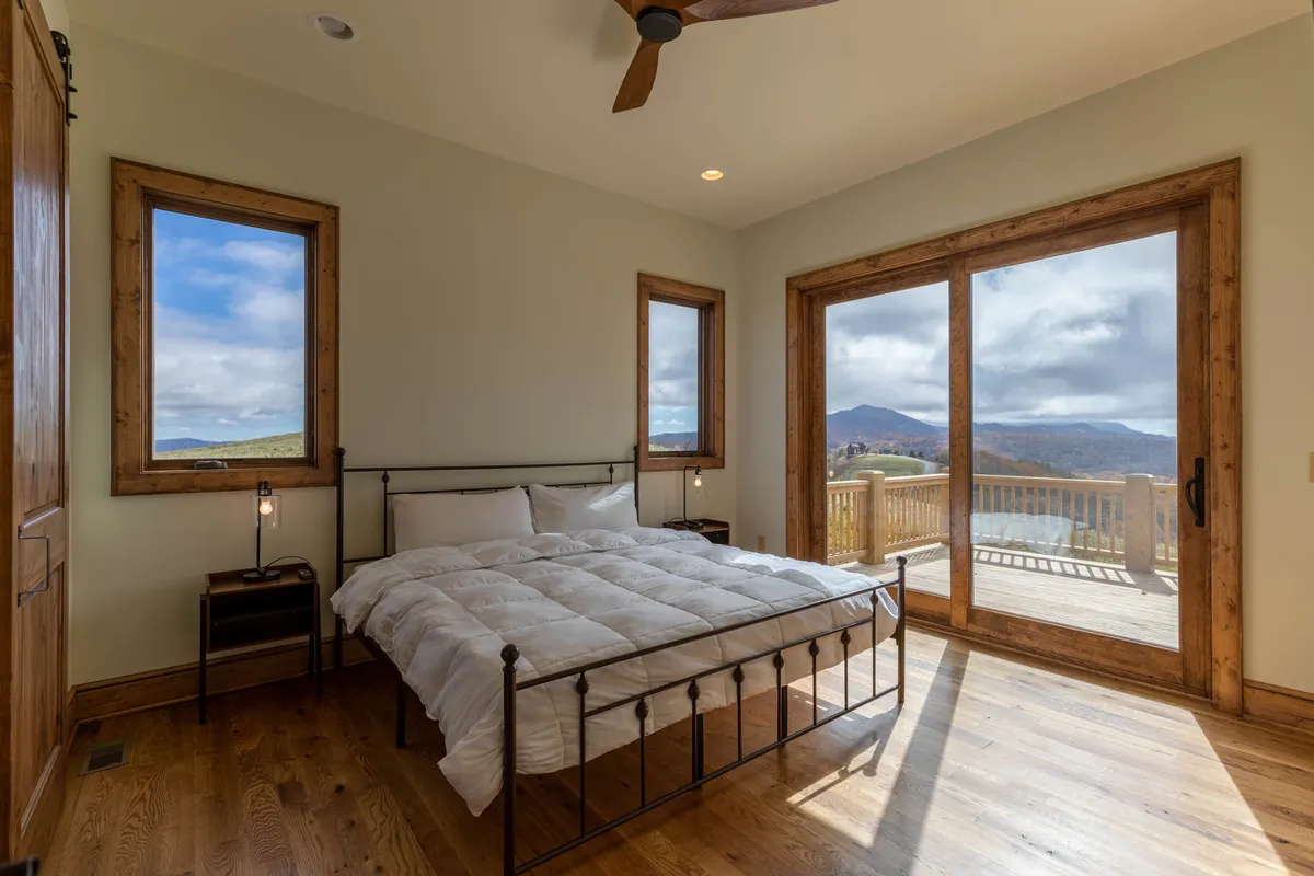 A serene bedroom with a comfortable bed facing large windows that let in abundant natural light, wooden flooring, and tasteful decor. The view through the windows reveals the beautiful mountain scenery, and a balcony is just beyond the doors, inviting guests to step outside.