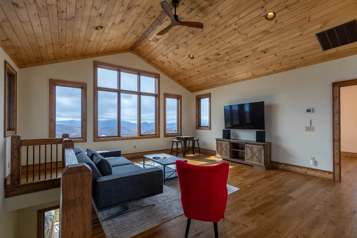 A cozy lounge space with a plush grey sofa, red accent chair, entertainment unit, and expansive windows peering out at the mountains. The wooden ceiling and homey decor make it an inviting hub for relaxation, games, or family gatherings.