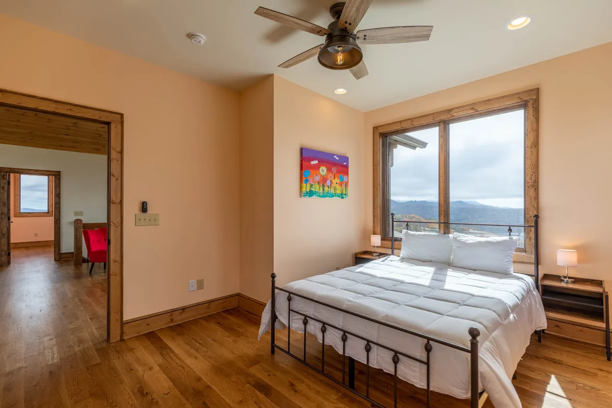 This image showcases a bed with crisp white bedding positioned beside large windows that open up to scenic mountain views. The colorful artwork on the wall adds vibrance, while natural light fills the space, creating a seamless connection between indoors and the outdoor surroundings.