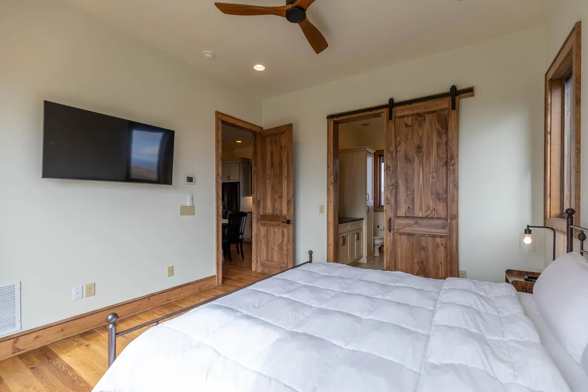 A serene suite with a plush bed dressed in white comforter, wooden sliding doors, and a mounted TV. The window lets natural light flood the room, creating a tranquil and comfortable atmosphere perfect for rest and privacy.