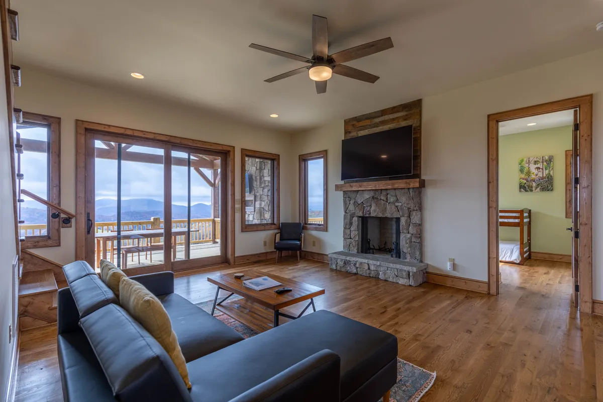 A modern, minimalist living room with a stone fireplace, sleek sofa, and a wooden coffee table, all oriented toward large windows that capture breathtaking mountain vistas. The room is filled with natural light, creating a warm, inviting hub for relaxation and socializing.