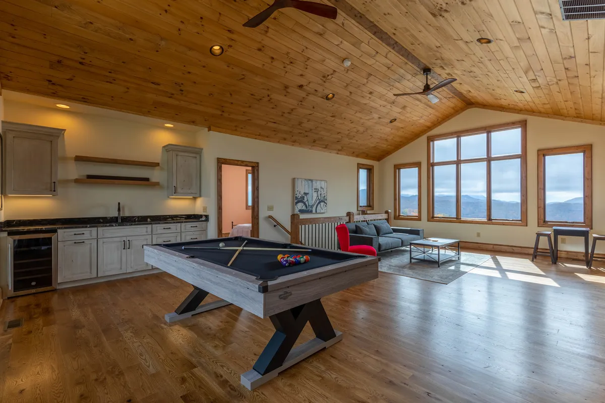 This image captures a game area that includes a pool table, comfortable sofa, kitchenette, and large windows displaying a scenic backdrop. The setup is ideal for both competitive games and relaxed movie nights, with easy access to snacks and refreshments.